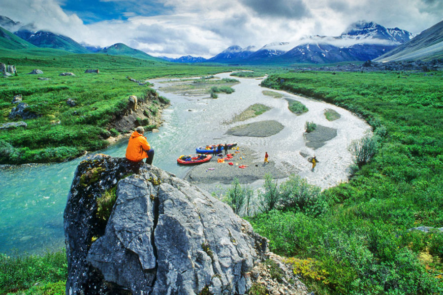 Man perched on rock high above the Snake River in Yukon Canada. Several rafts and canoes can be seen on the river below.