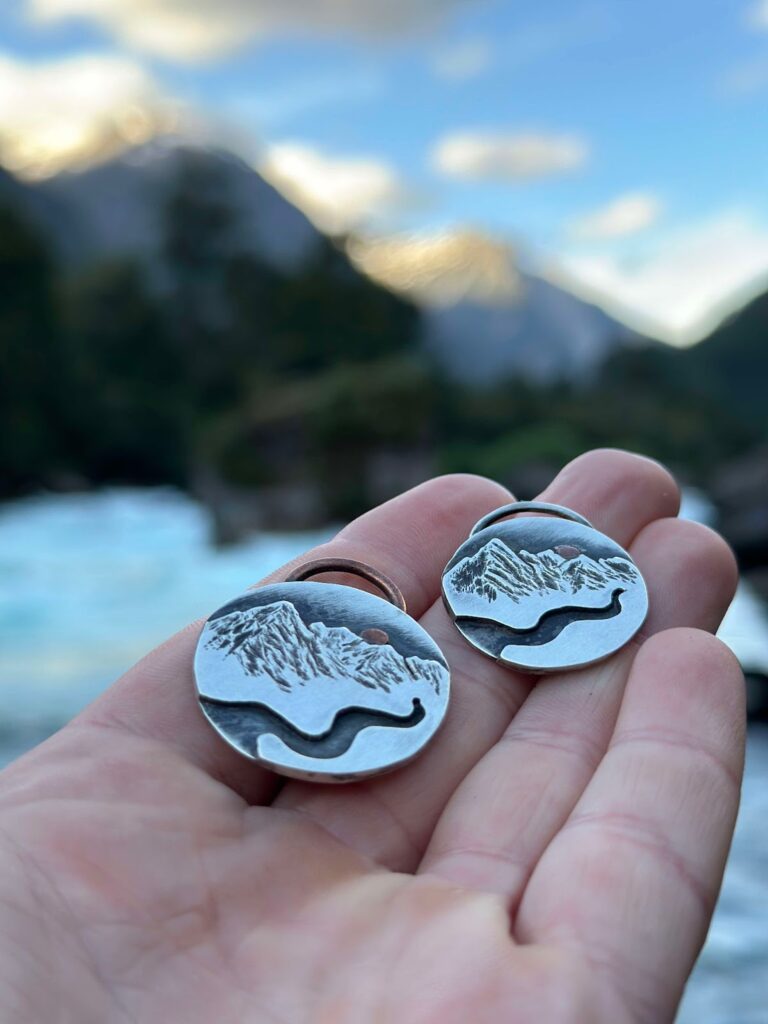 a hand holding a pair of silver earrings with mountains in the background
